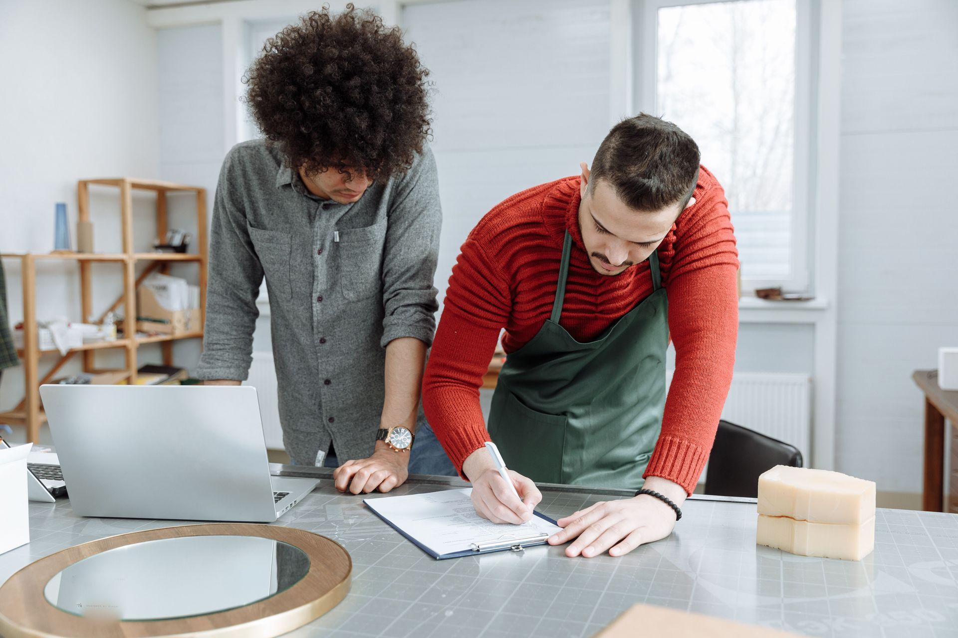 Two people collaborating over plans at a worktable in a bright studio, with a laptop and tools nearby