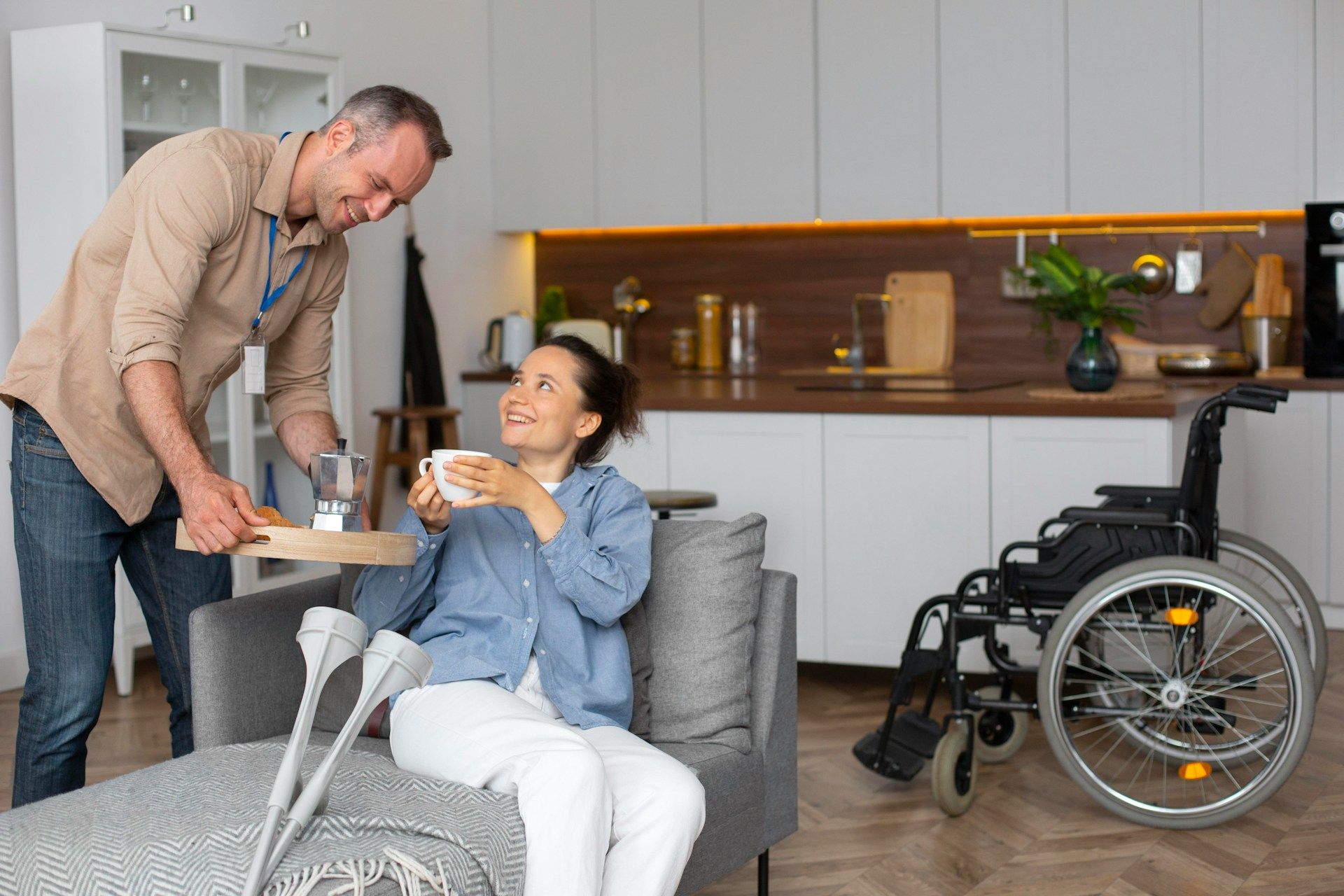 Caregiver serving a drink to a smiling woman in a wheelchair in a modern living room