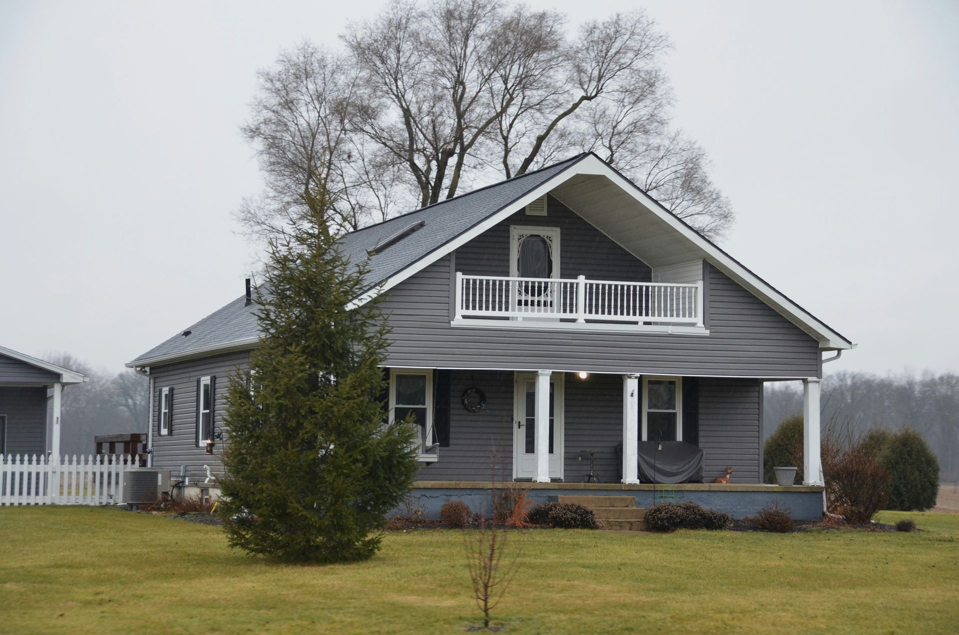 Gray house with white porch and balcony on a foggy lawn, with bare trees behind it