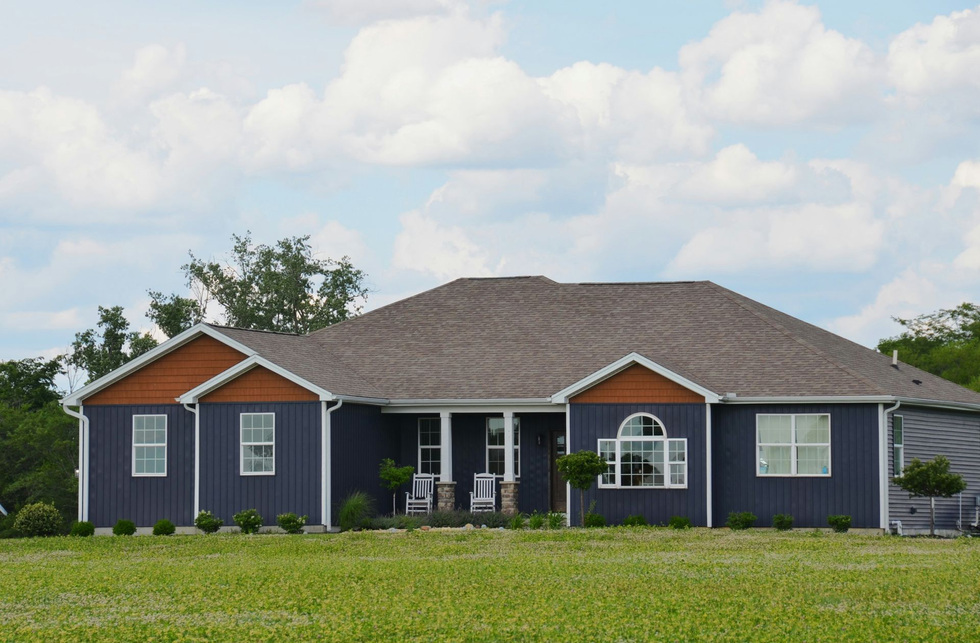 Single-story blue-gray house with a dark roof and white trim, set in a grassy yard under a cloudy sky