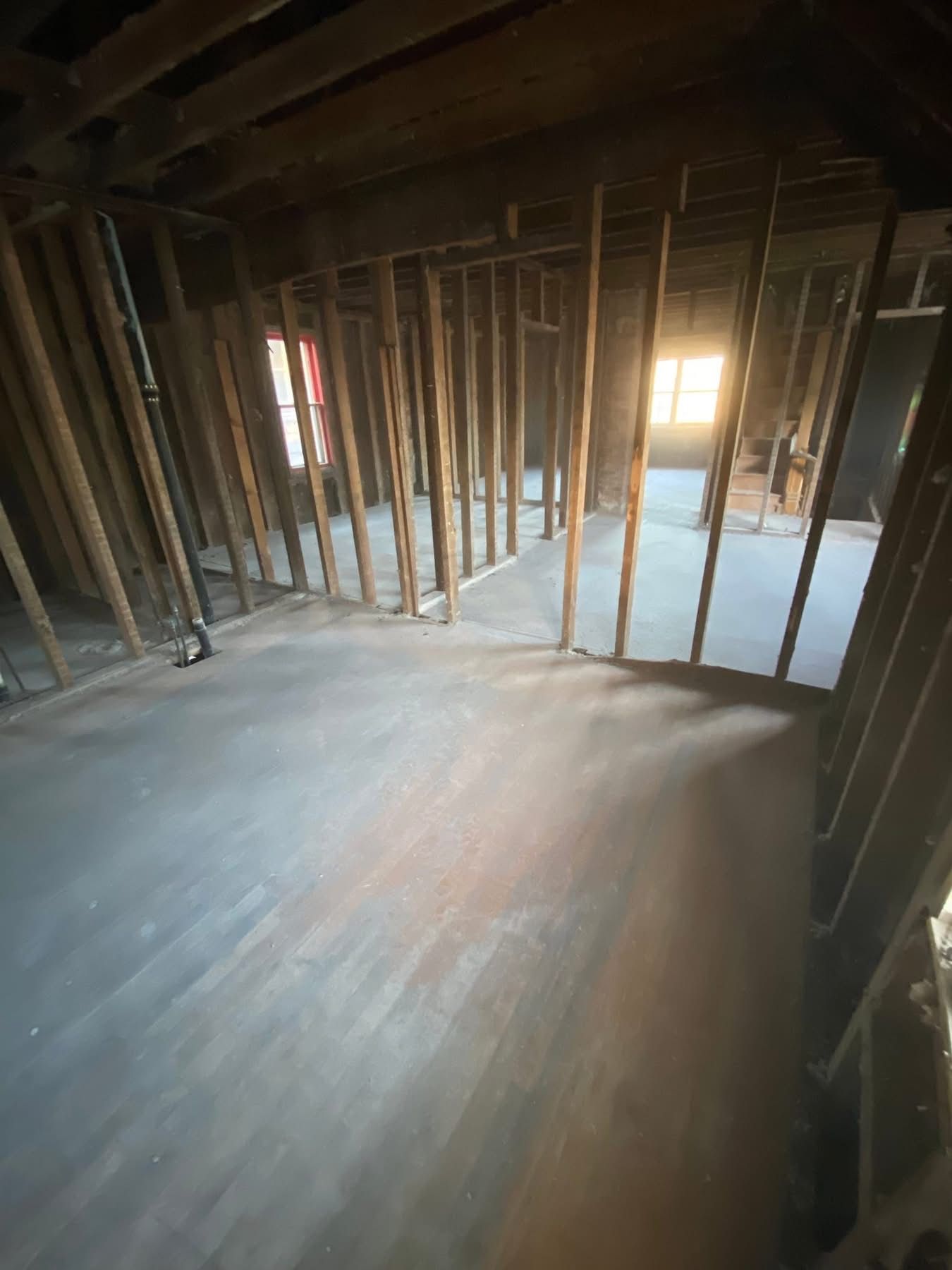 Interior of a room under construction, with exposed wooden beams and floorboards. Sunlight streams in from a window.
