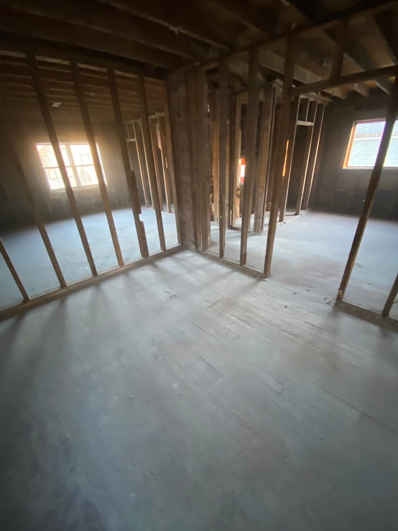 Interior of a room under construction, with wooden studs and floorboards. Sunlight streams through windows.