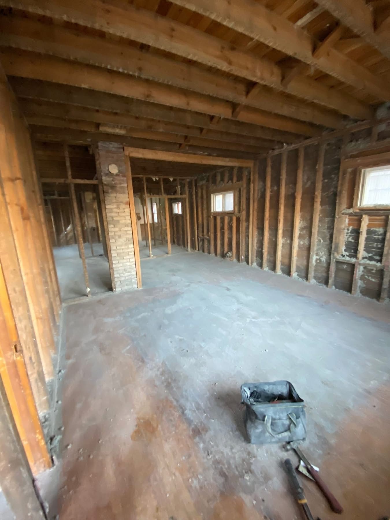 Interior of a room under construction, with exposed wooden studs, beams, and a brick chimney. A toolbox rests on the floor.