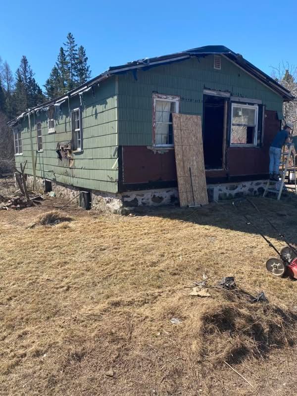 Old house with damaged siding and exposed foundation, in a grassy area. One person stands nearby.