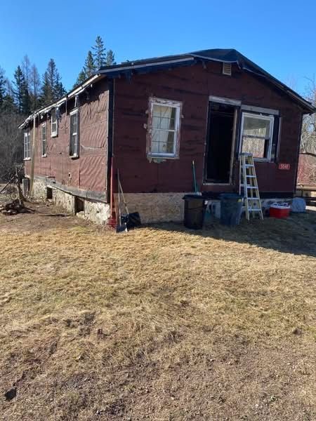 Dilapidated red building with open doorway, ladder, and trash. Dry grass in front, blue sky background.