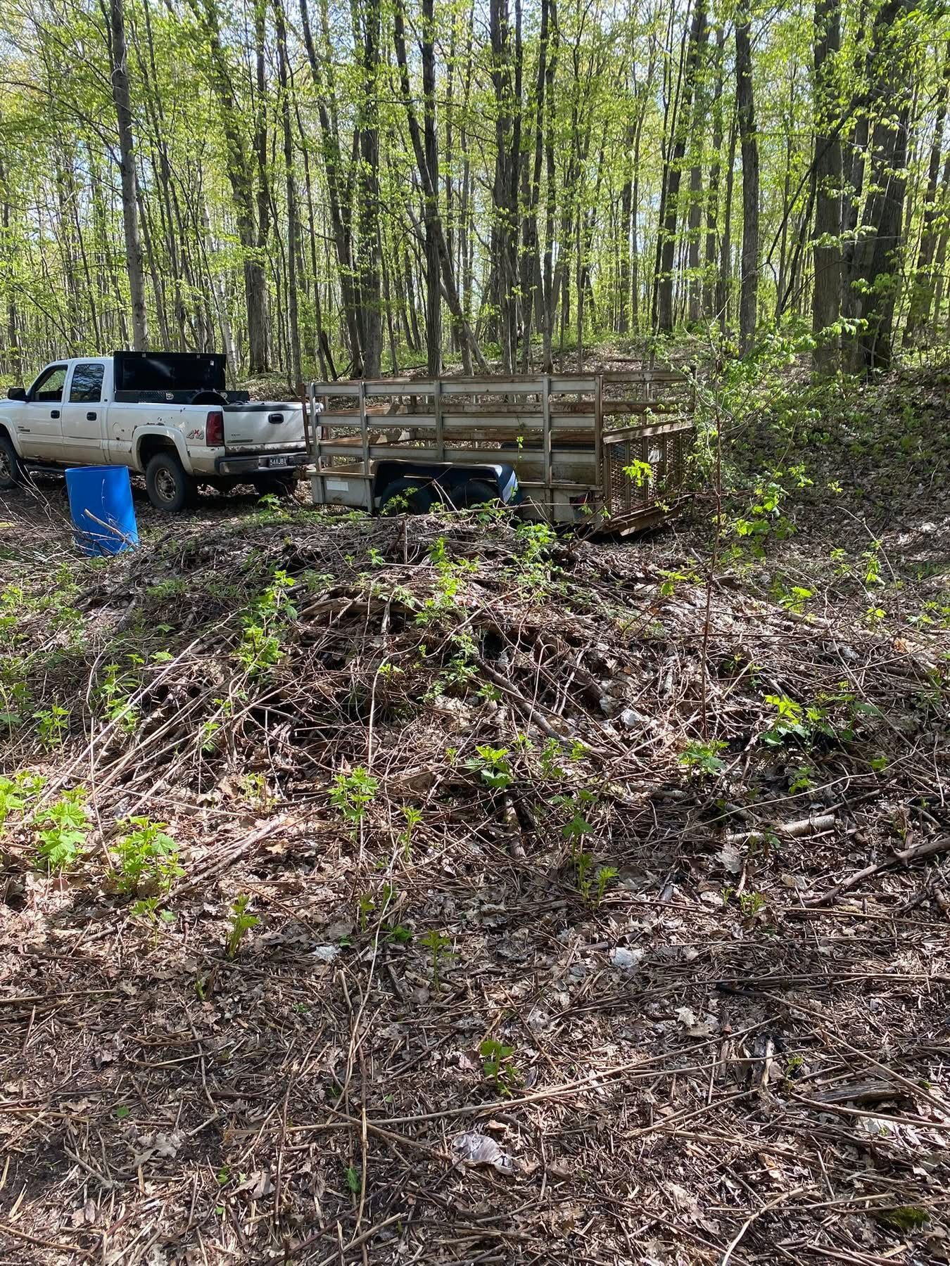 White pickup truck and trailer parked near a large pile of leaves in a wooded area.