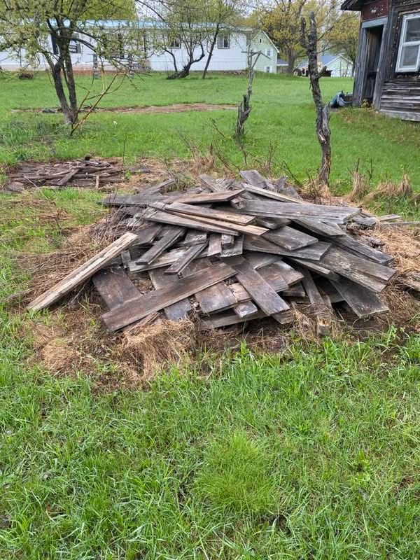 Pile of weathered wooden planks on grass, beside a dilapidated building.