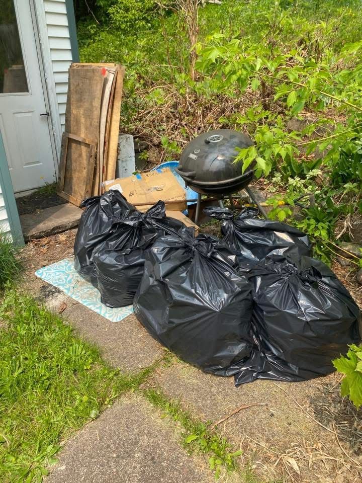 Four black trash bags on a concrete pad, next to a grill and debris near a white door and greenery.