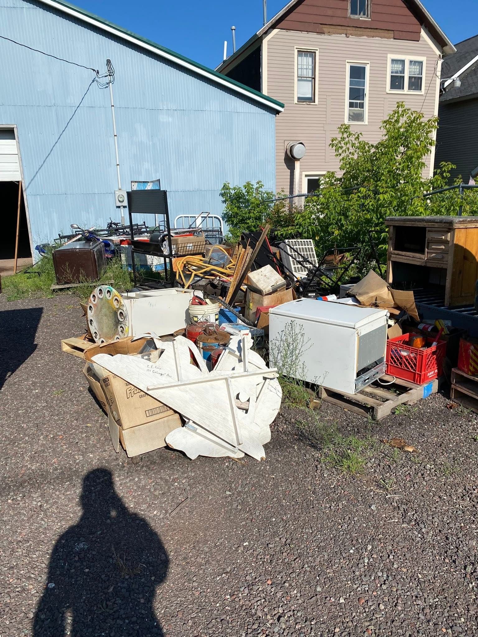 Pile of discarded items on gravel in front of a blue building, with a house in the background.