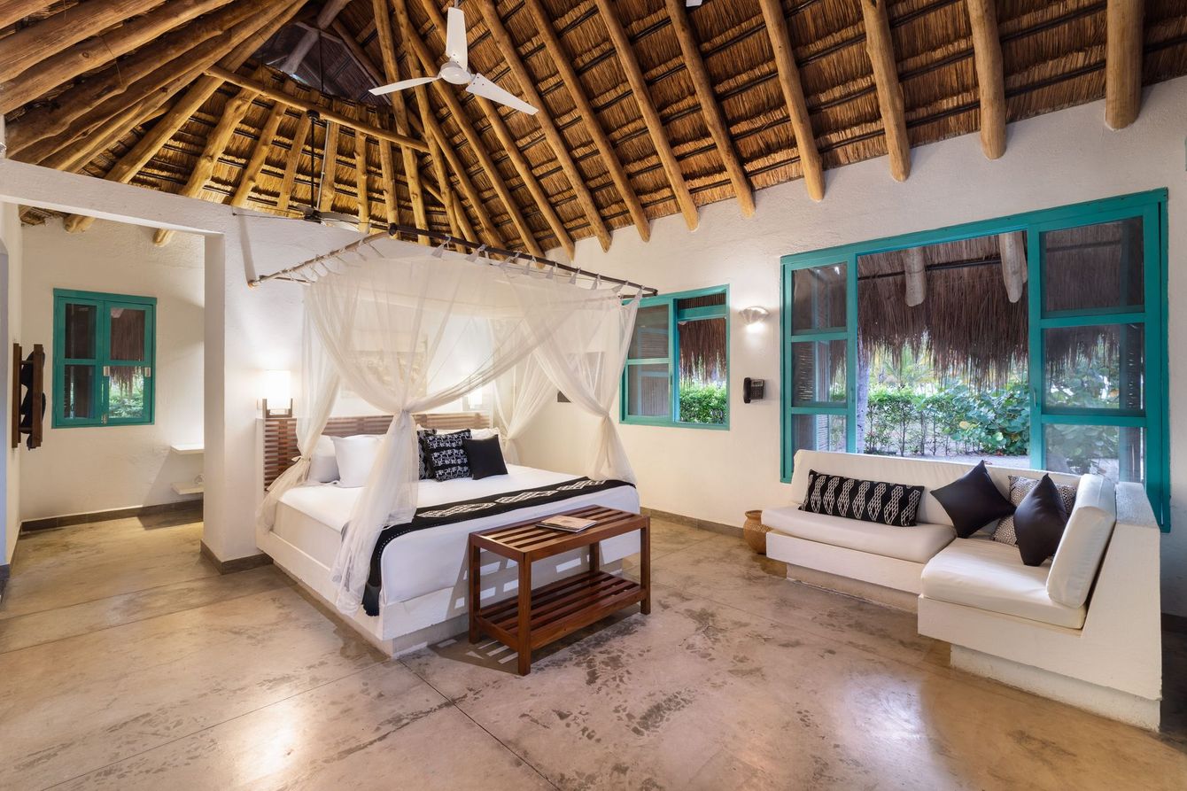 Habitación tropical con techo de paja, cama blanca y sofá a juego junto a ventanas de color turquesa brillante.