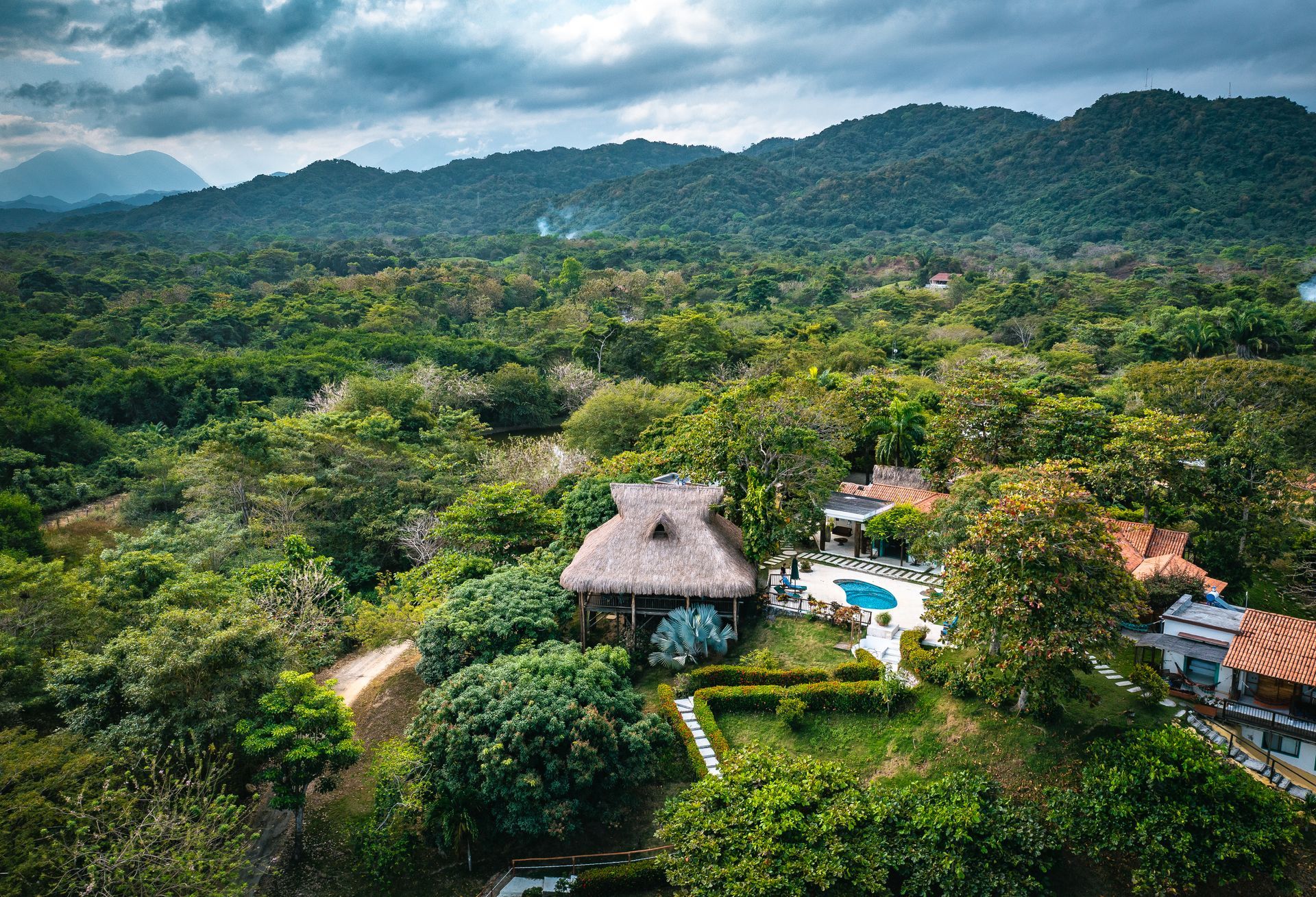 Vista aérea de un complejo turístico tropical en la ladera de una colina, con cabañas de techo de paja, una piscina y un denso bosque verde.