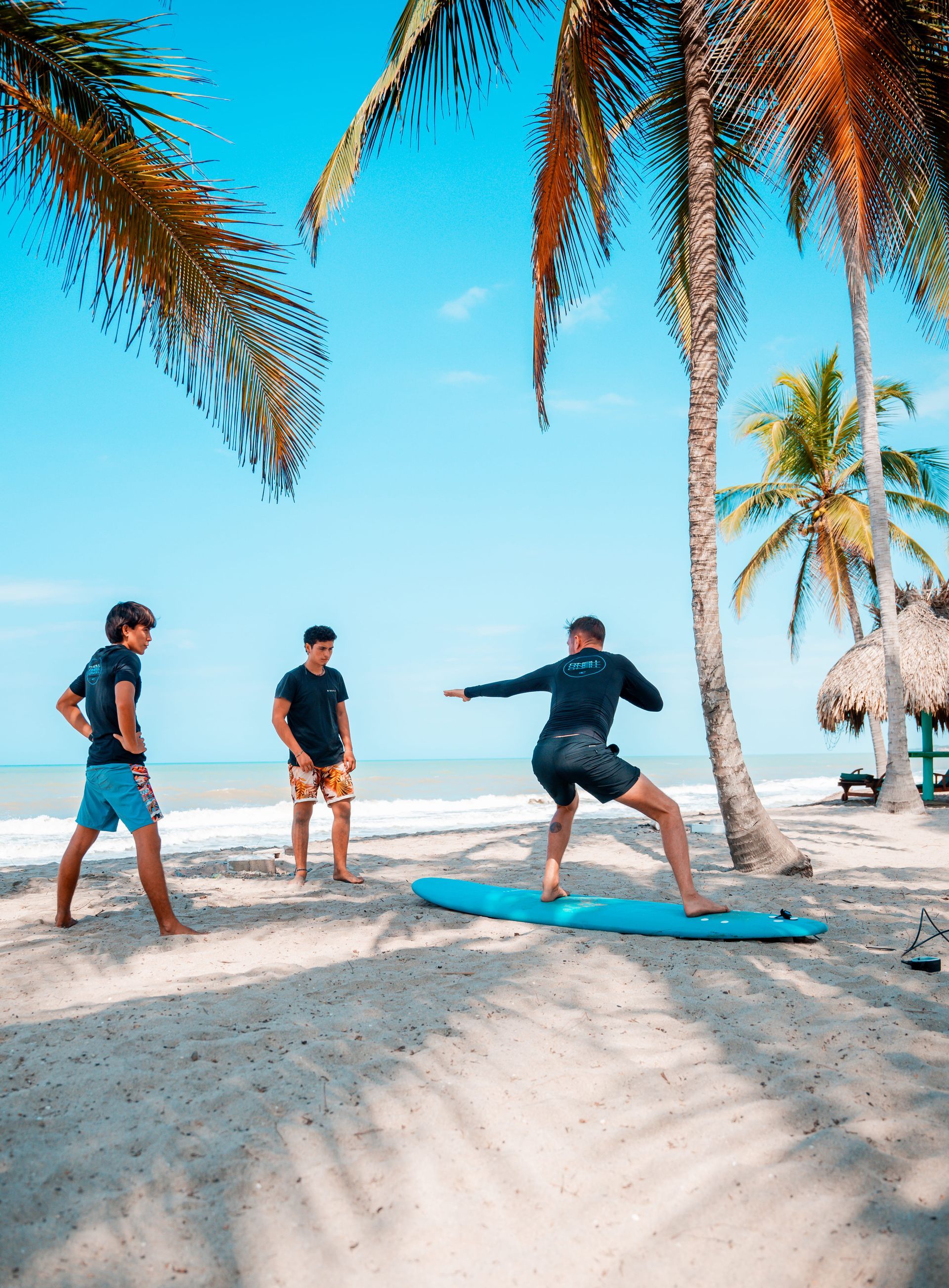 Tres personas en una playa, una de ellas haciendo equilibrio sobre una tabla de surf azul bajo unas palmeras.