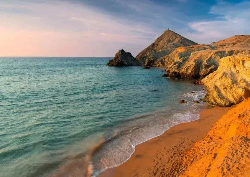 Playa de arena con aguas turquesas, acantilados rocosos y un cielo rosado al atardecer.