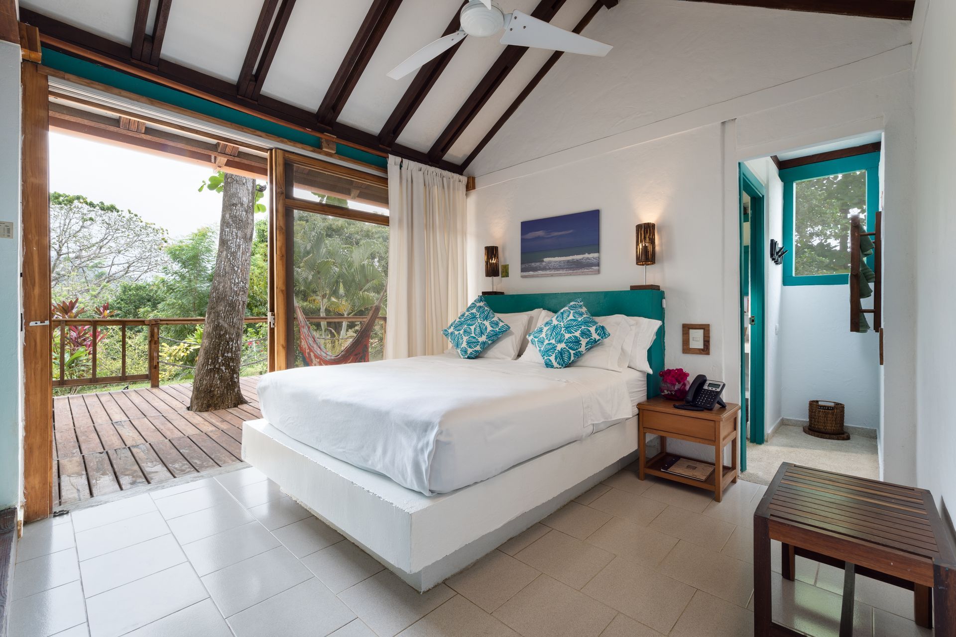 Habitación tropical con cama blanca, almohadas color turquesa, terraza abierta y mucha luz natural.