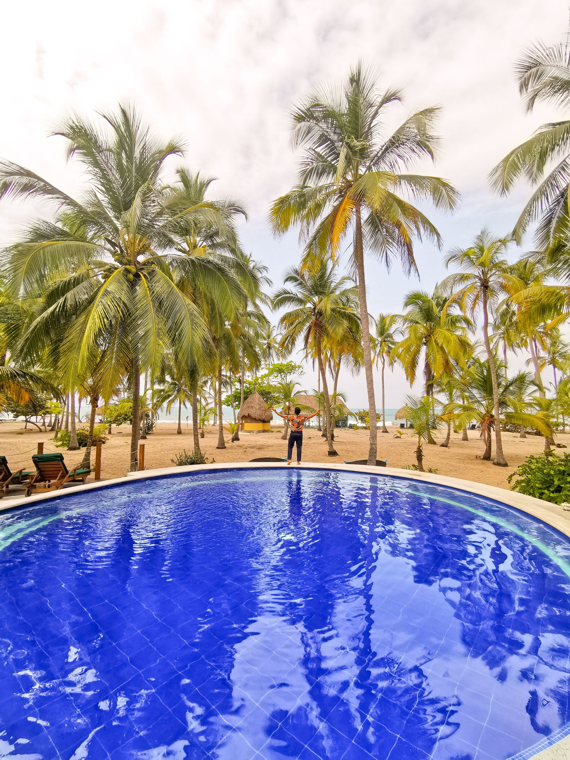 Piscina azul con palmeras y una playa de arena al fondo bajo un cielo brillante.
