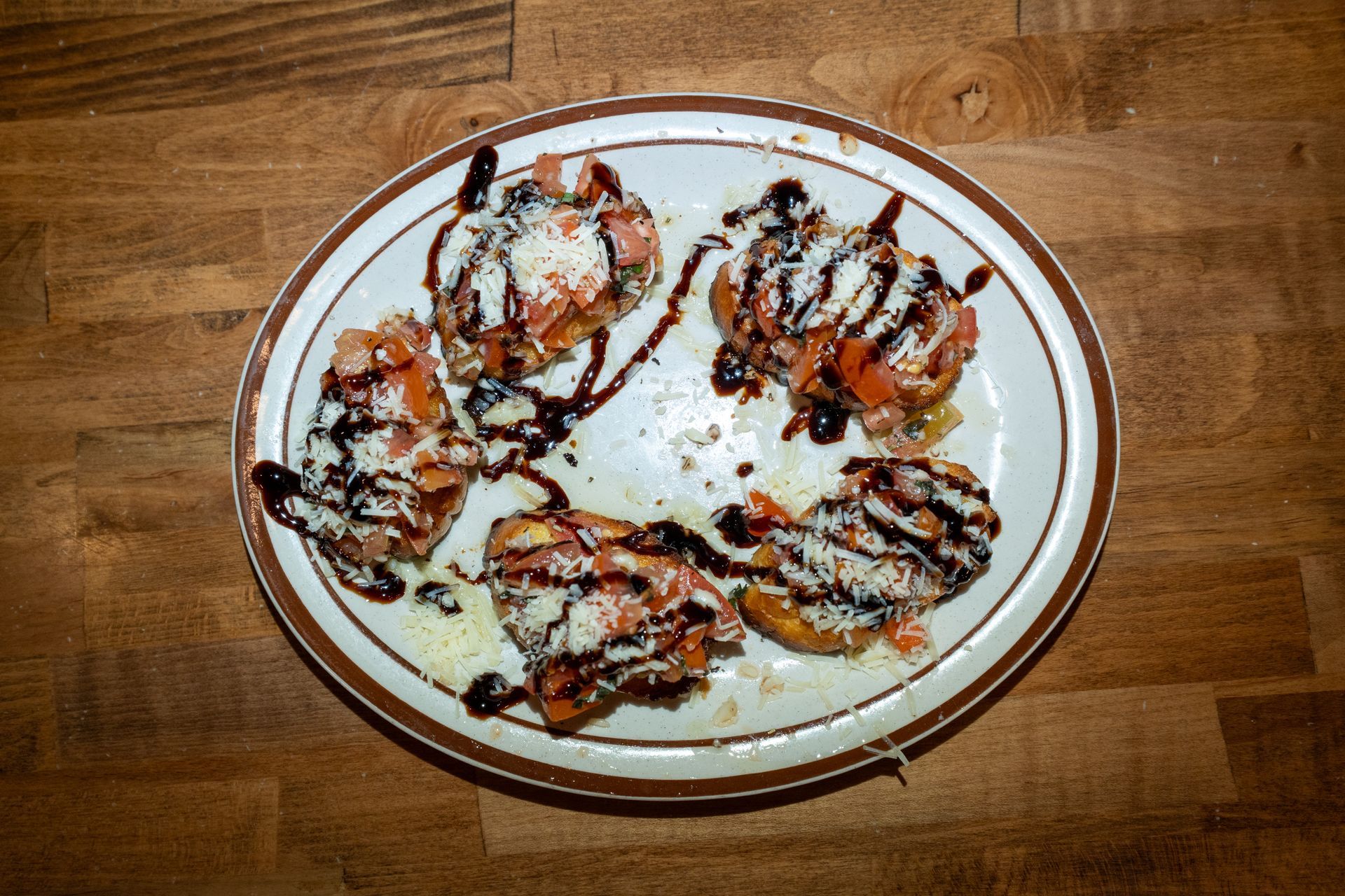 Bruschetta appetizer on a plate, drizzled with balsamic glaze and grated cheese, on a wooden surface.