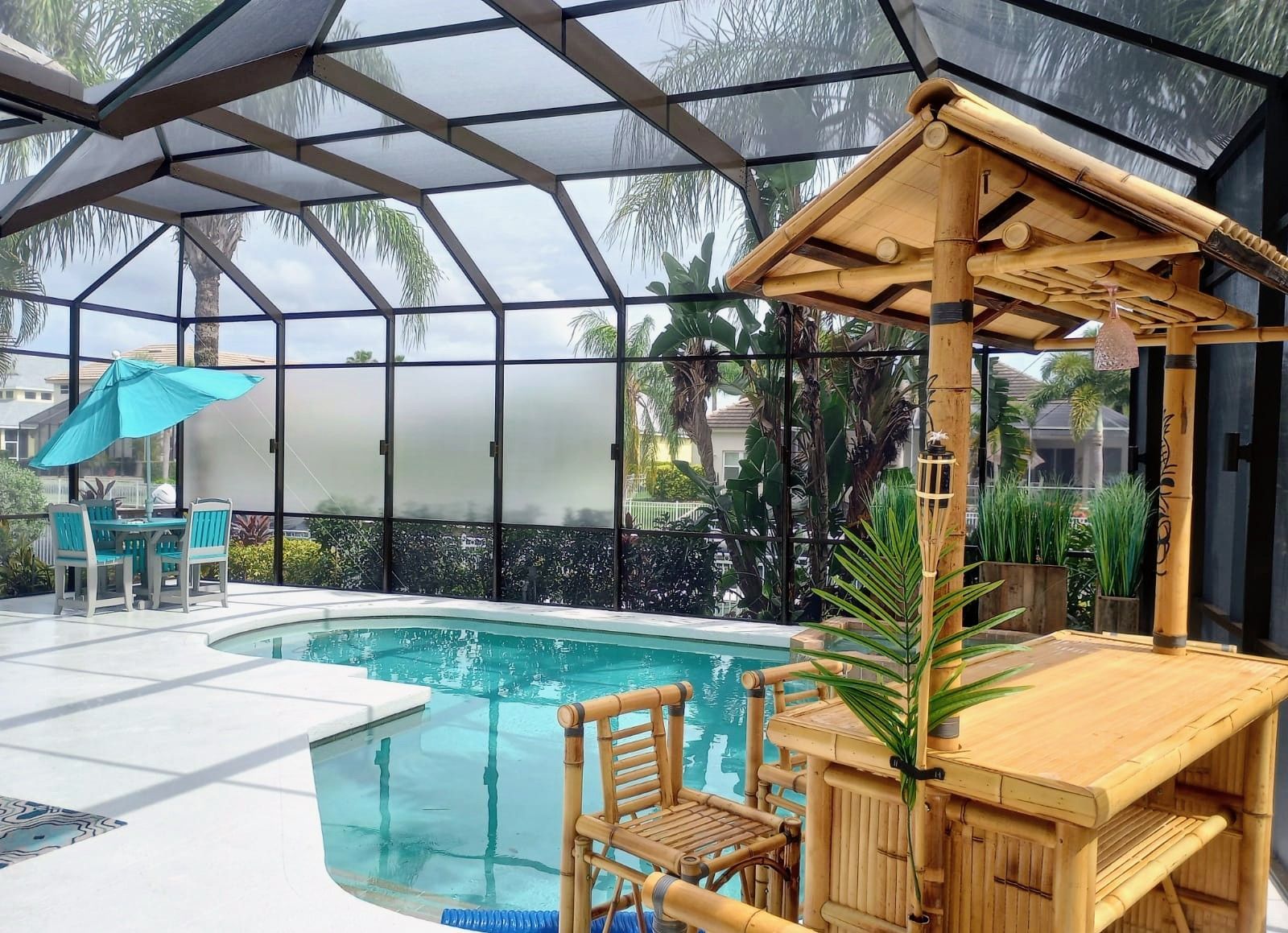 A swimming pool inside a screened-in lanai with a bamboo tiki bar and a small table with chairs under a blue umbrella.