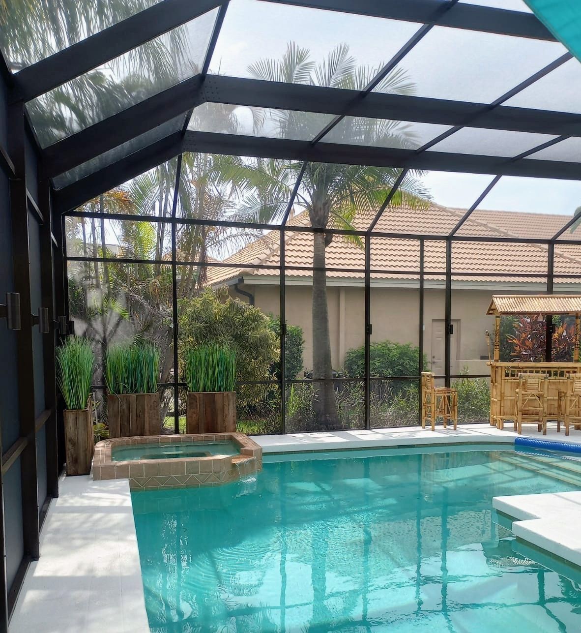 A screened-in patio featuring a swimming pool, a hot tub, potted plants, and a tiki bar area.