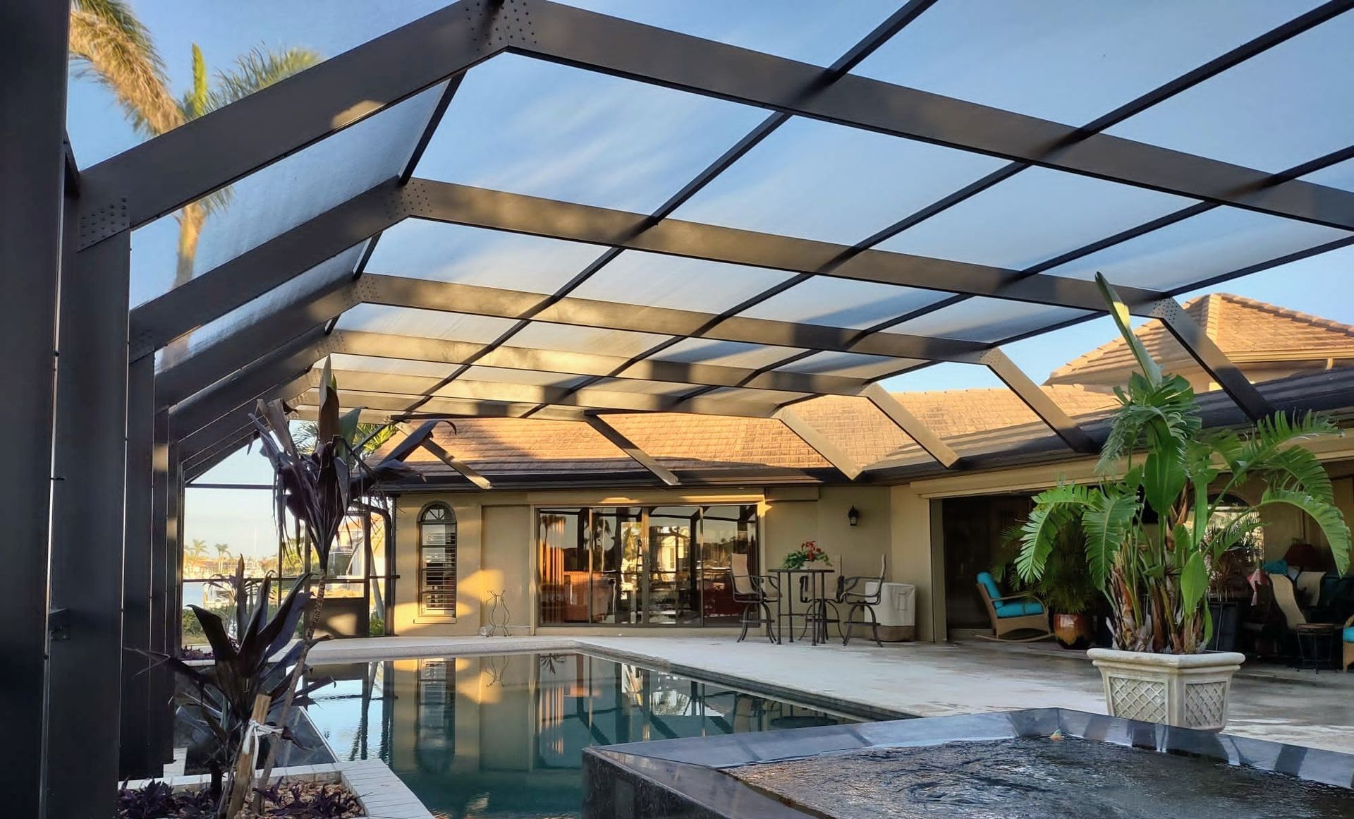 A sunlit backyard swimming pool under a large, dark metal-framed screen enclosure next to a house patio.