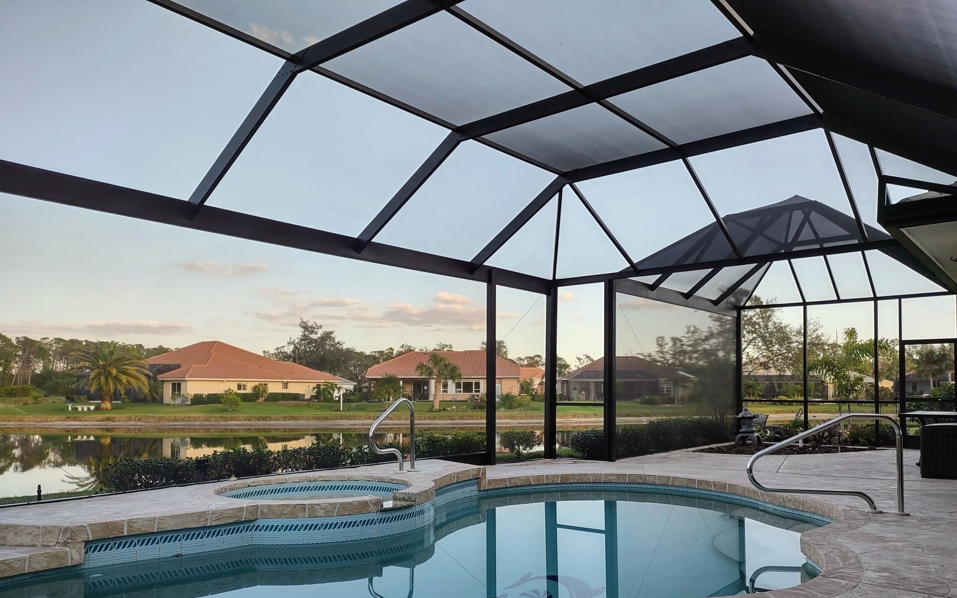 A screened-in swimming pool and spa overlooking a lake surrounded by suburban homes at sunset.