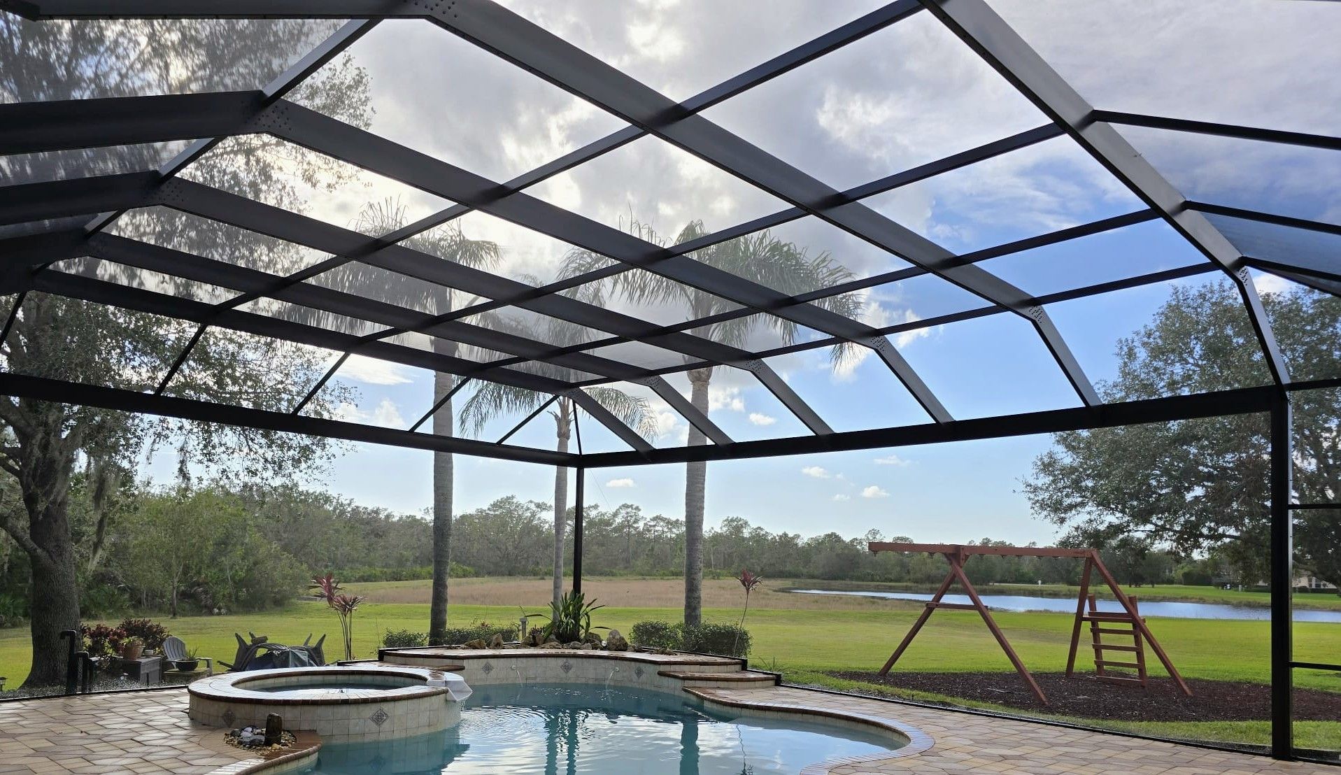 A screened-in patio overlooking a backyard pool, a small wooden playset, a grassy lawn, and a distant pond under a blue sky.