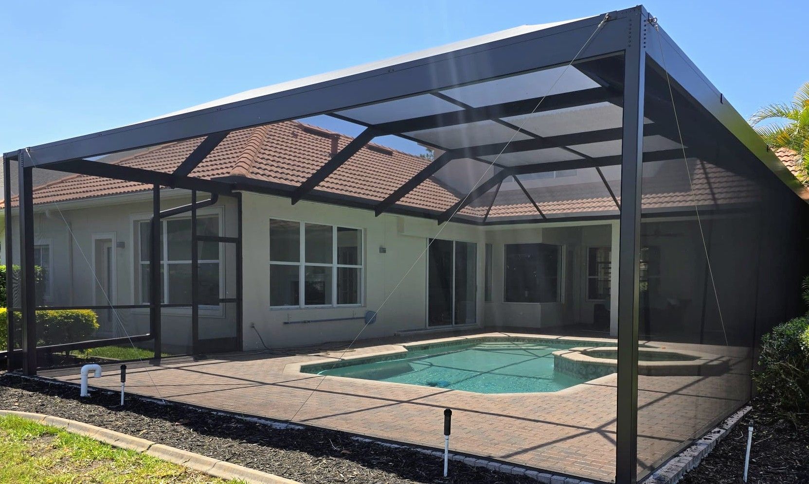Backyard pool with a rectangular light-colored patio, enclosed by a large black metal screen cage under a clear blue sky.
