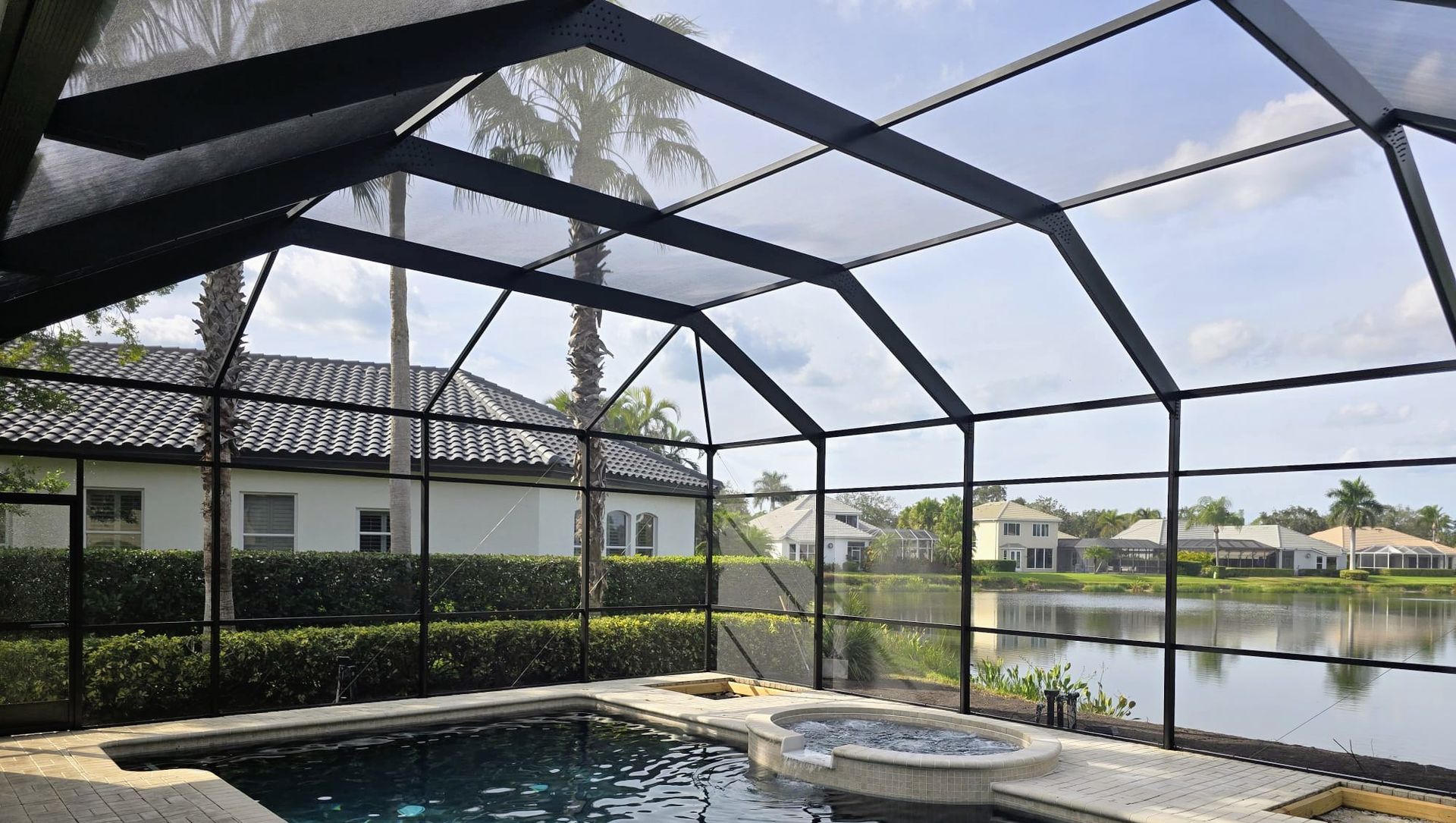 A swimming pool and hot tub enclosed within a black metal screen cage, overlooking a lake and houses.