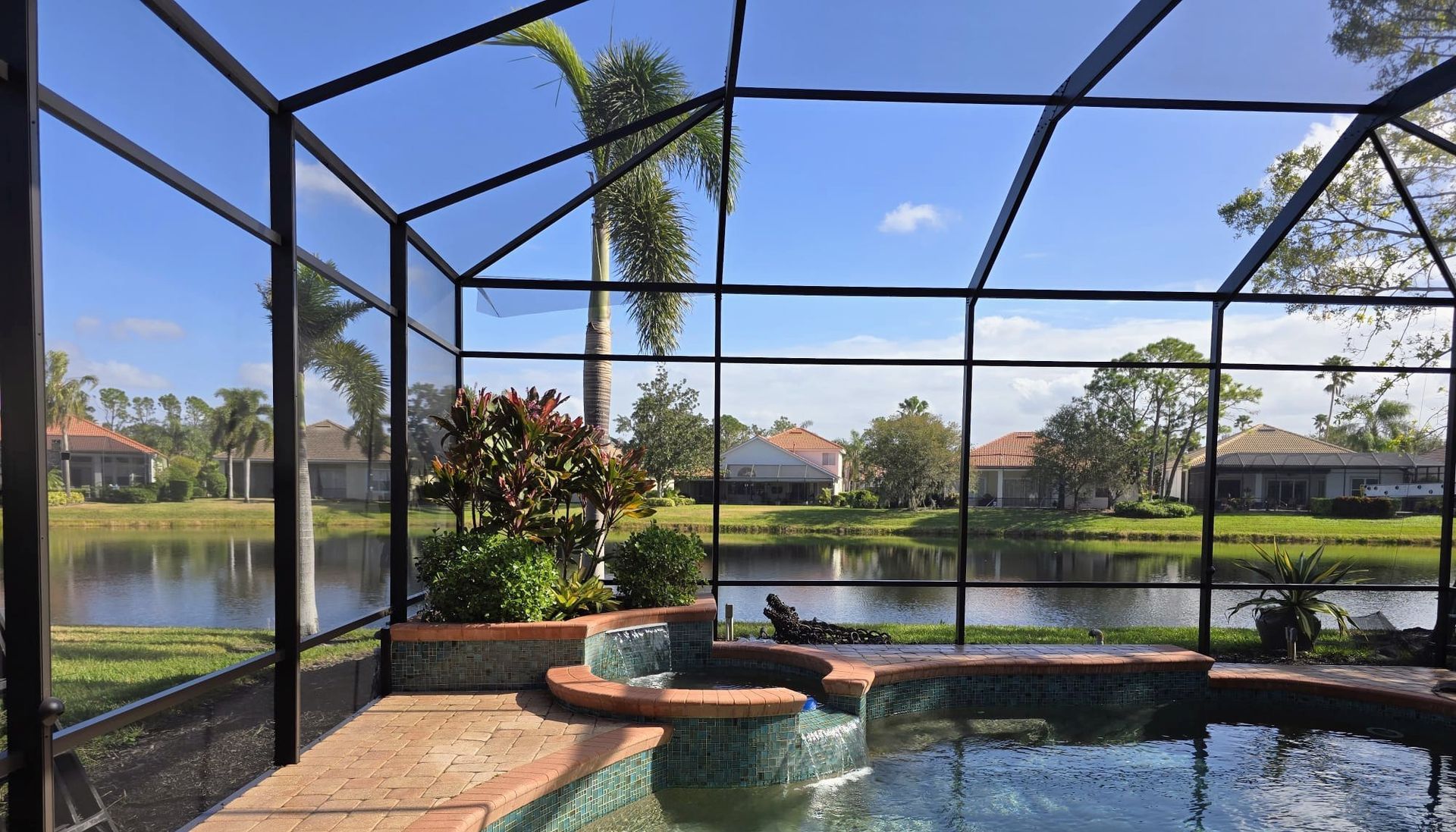 A view from inside a screened pool enclosure looking out at a backyard pond, palm trees, and neighborhood houses.