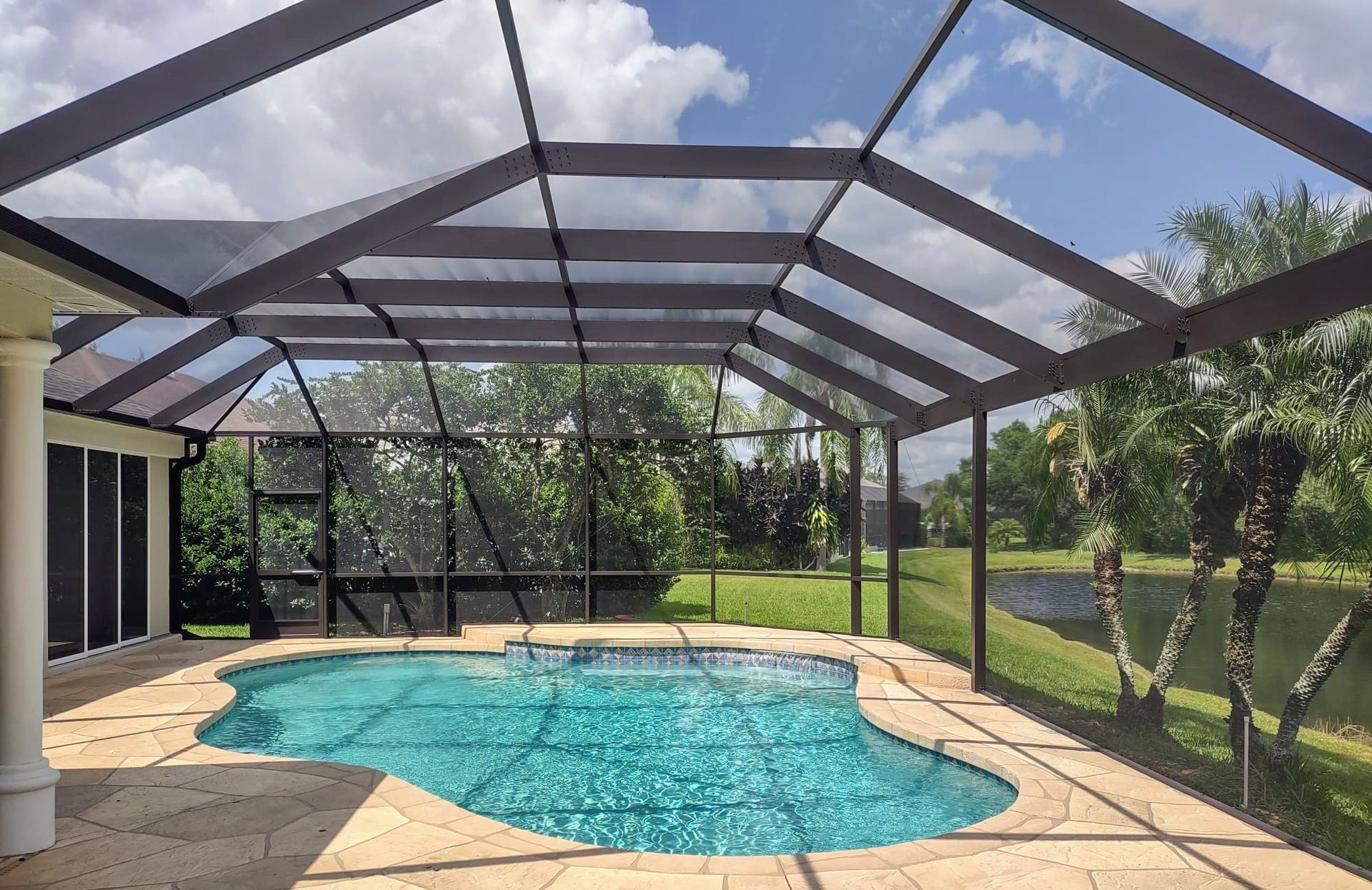 A backyard pool with a light blue surface, enclosed by a large screen cage on a tan stone deck overlooking a pond.