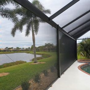 A patio enclosed by a dark screen overlooks a grassy lawn, a pond, and palm trees under a cloudy sky.