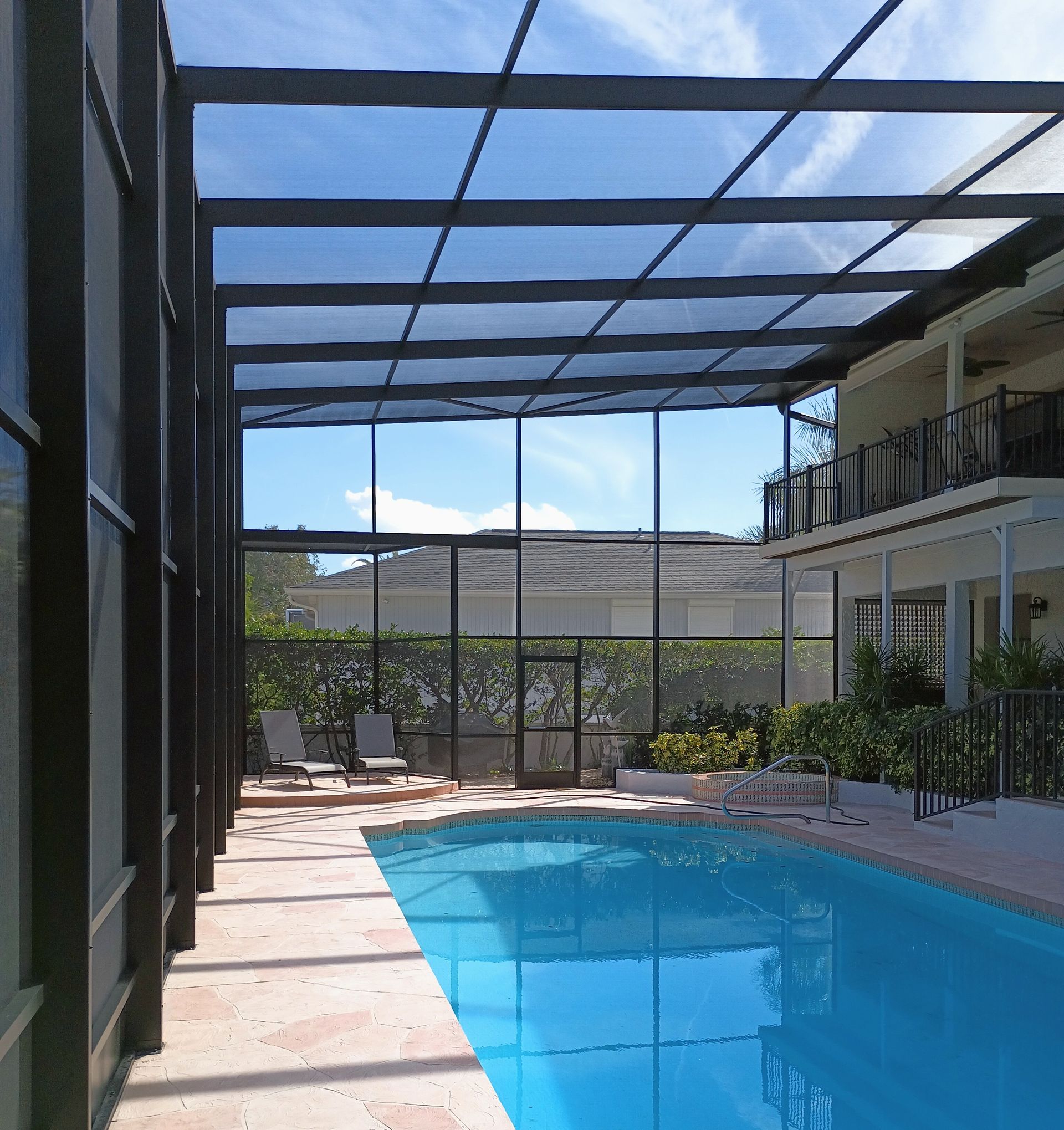 A swimming pool enclosed by a black metal frame and screen, with lounge chairs on a stone deck under a sunny blue sky.