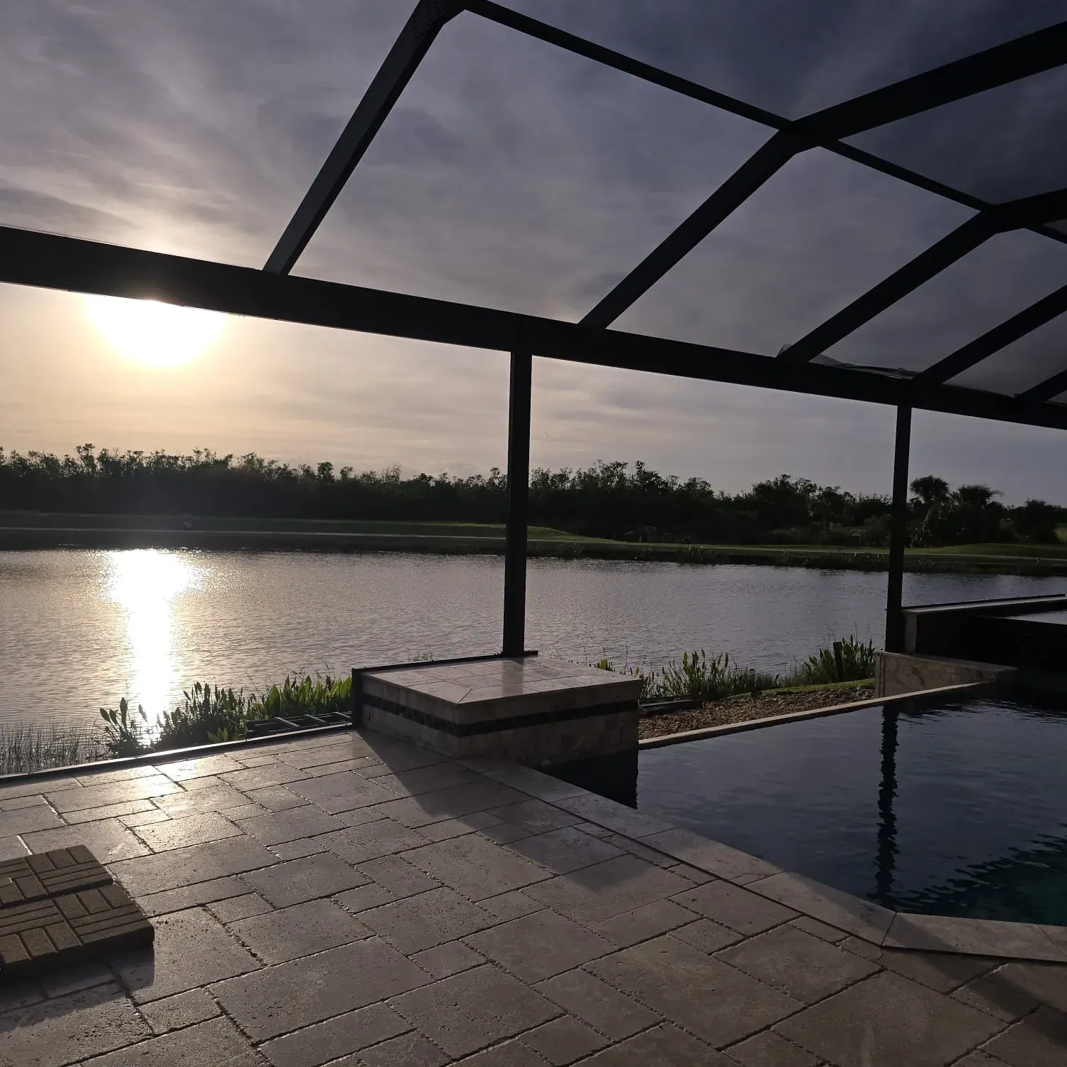 A sunset reflects off a lake beyond a stone patio and infinity pool enclosed by a screen frame.