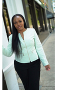 Woman standing outdoors in a light green jacket, with a blurred building and greenery behind her.