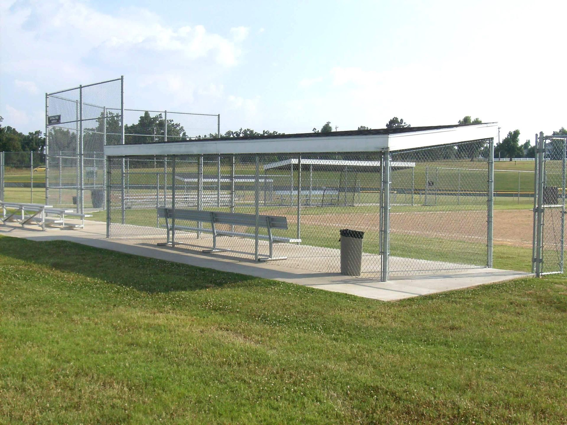 A baseball dugout with a metal roof, benches, and a trash can, situated on a grassy field beside a chain-link fence.