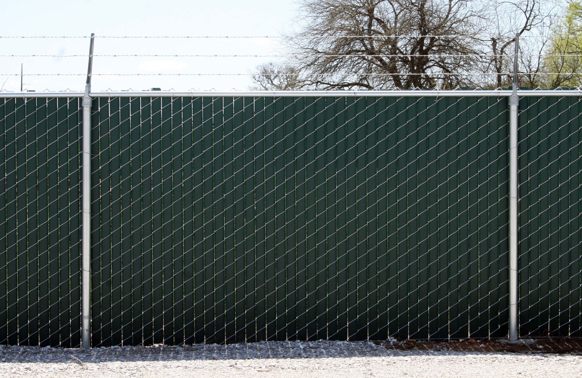 A green chain-link privacy fence with vertical slats and barbed wire on top, viewed from an outdoor gravel area.