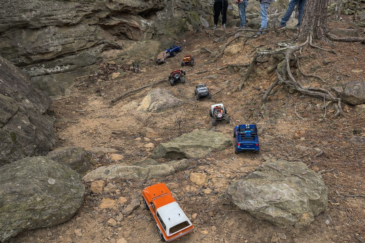 Three RC trucks driving over rocks with people watching outdoors.