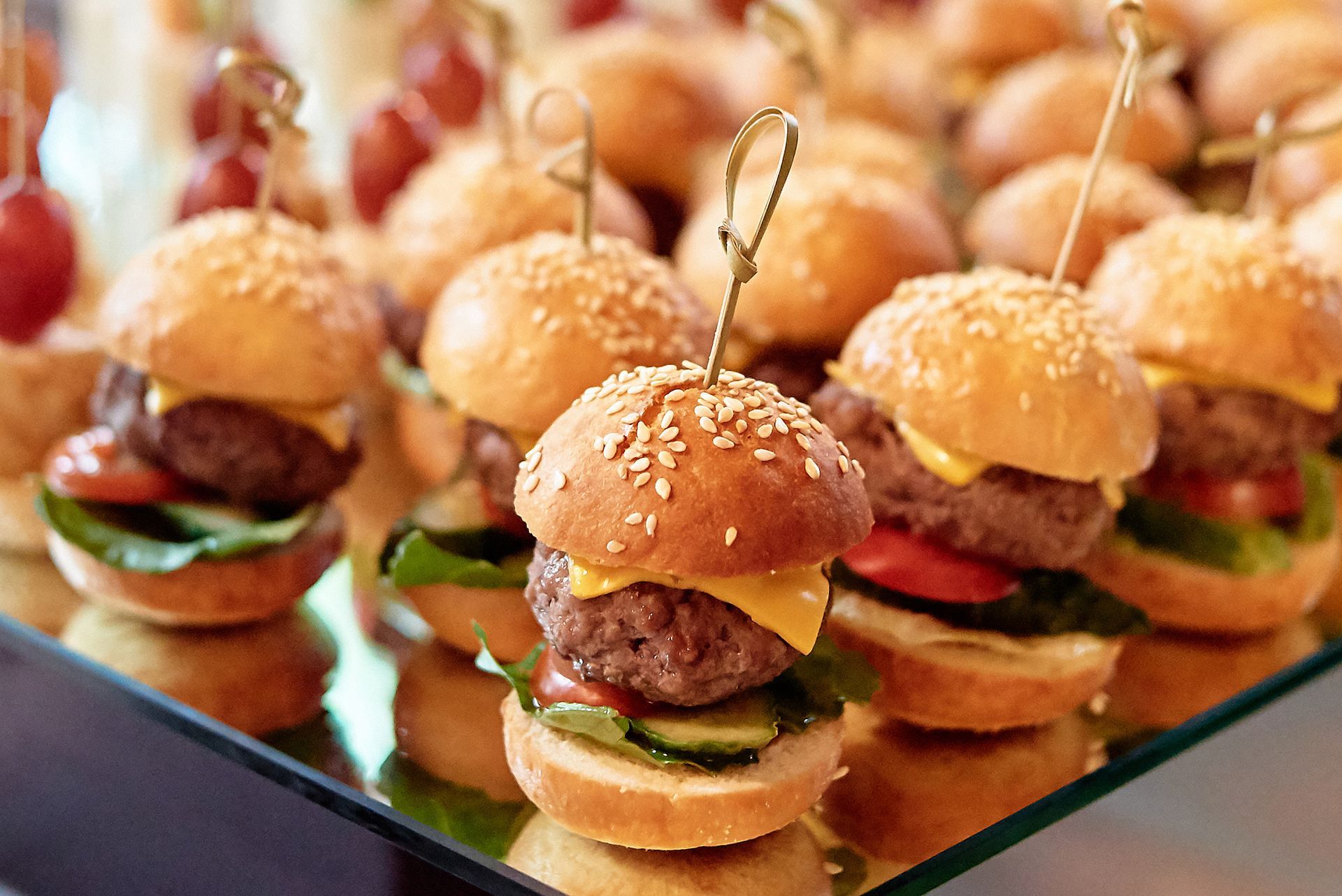 Mini burgers with sesame seed buns on a mirrored surface.
