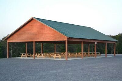 Wooden picnic pavilion with green roof and picnic tables on a gravel lot.