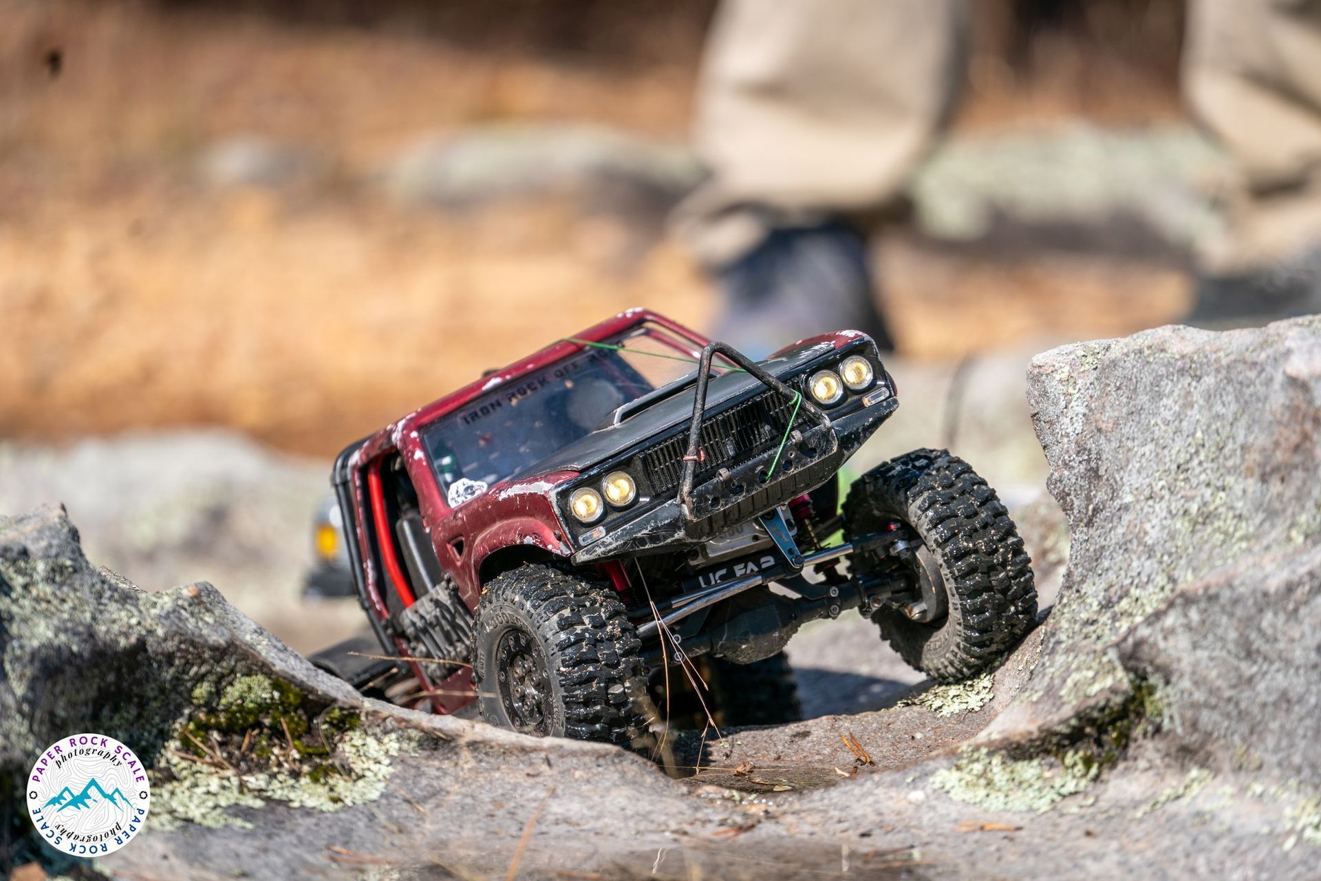 Small red remote-controlled truck climbing over rocks on a course; black tires, outdoor setting.