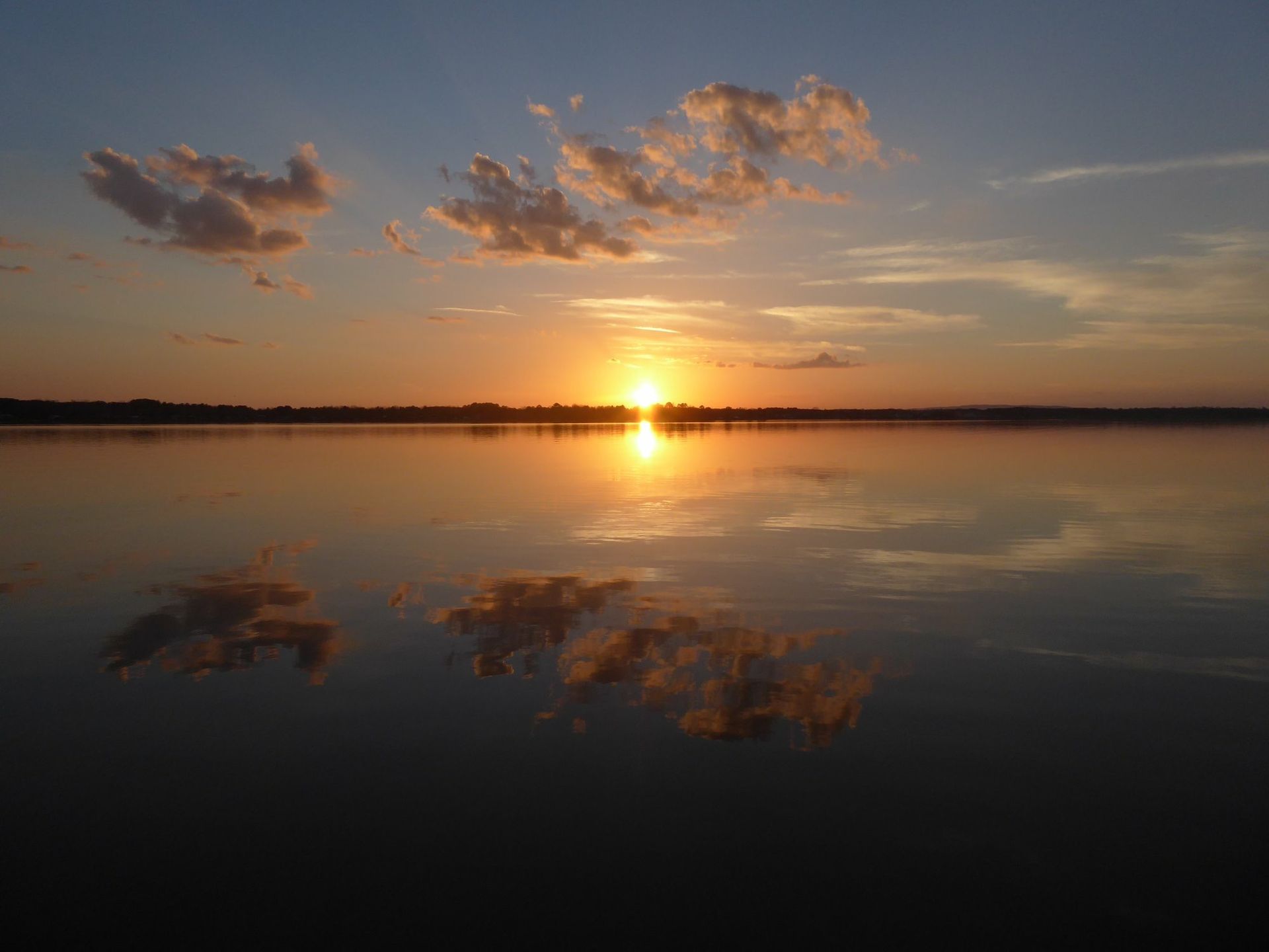 Sunset over a calm lake, reflecting orange sun and fluffy clouds.