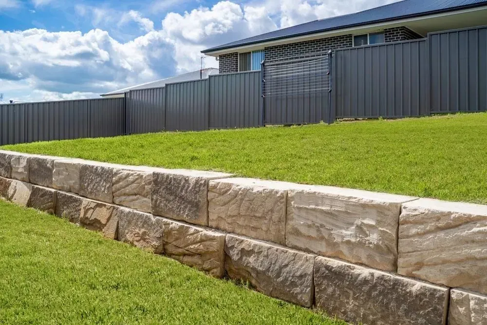 A Sandstone Retaining Wall Separates Two Levels of Green Lawn in A Yard — Creative Landscapes Coffs Harbour in Sawtell, NSW