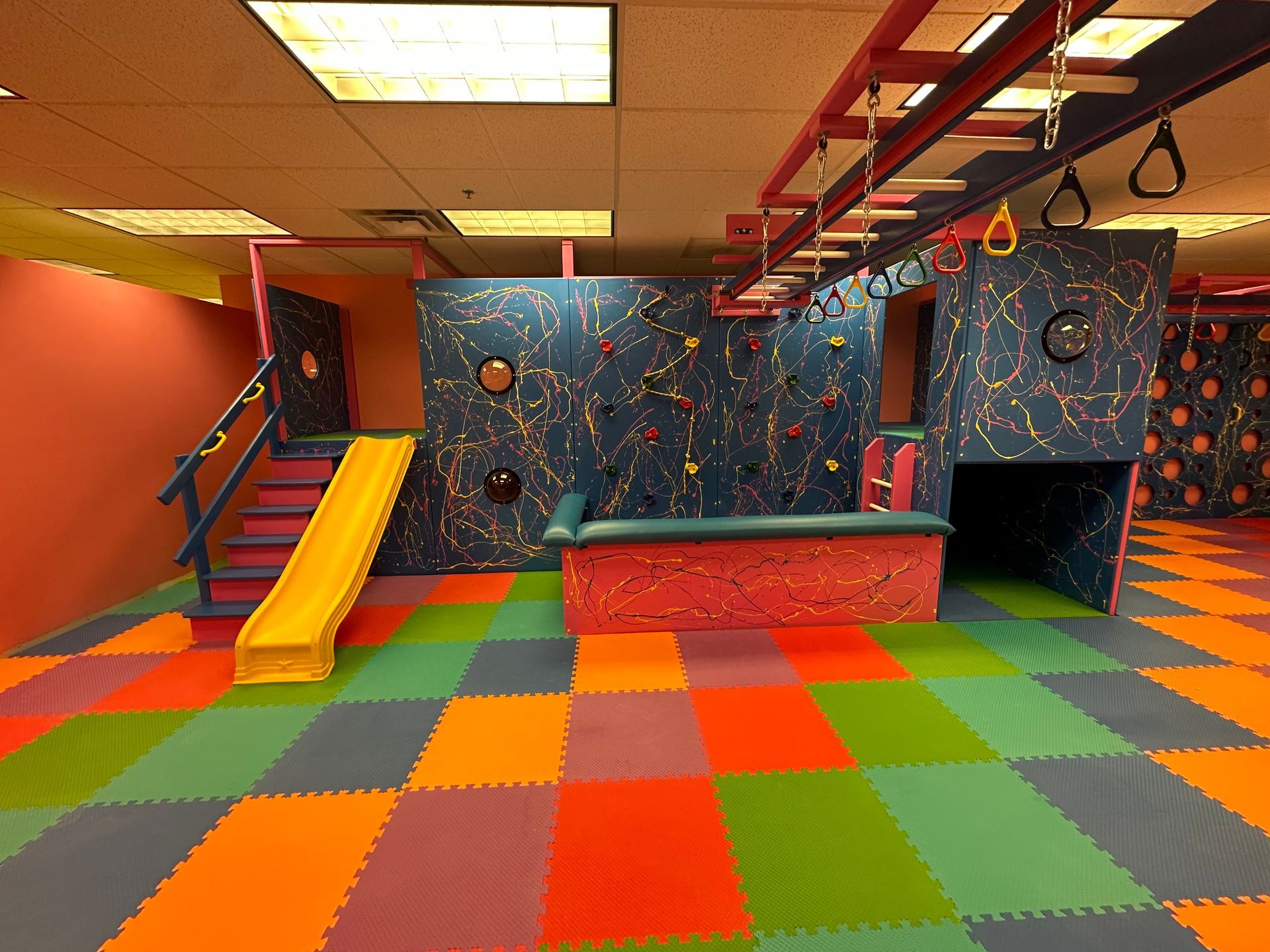 Commercial sensory gym with slide, rock climbing wall, monkey bars, and colorful padded flooring