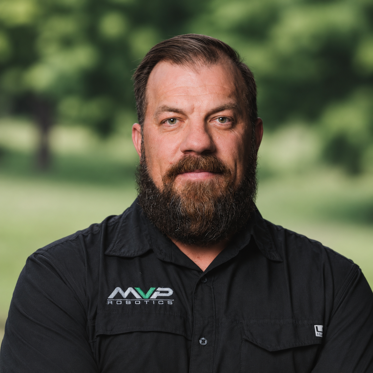 Portrait of a bearded man in a black shirt with a logo, standing outdoors against a blurred green background
