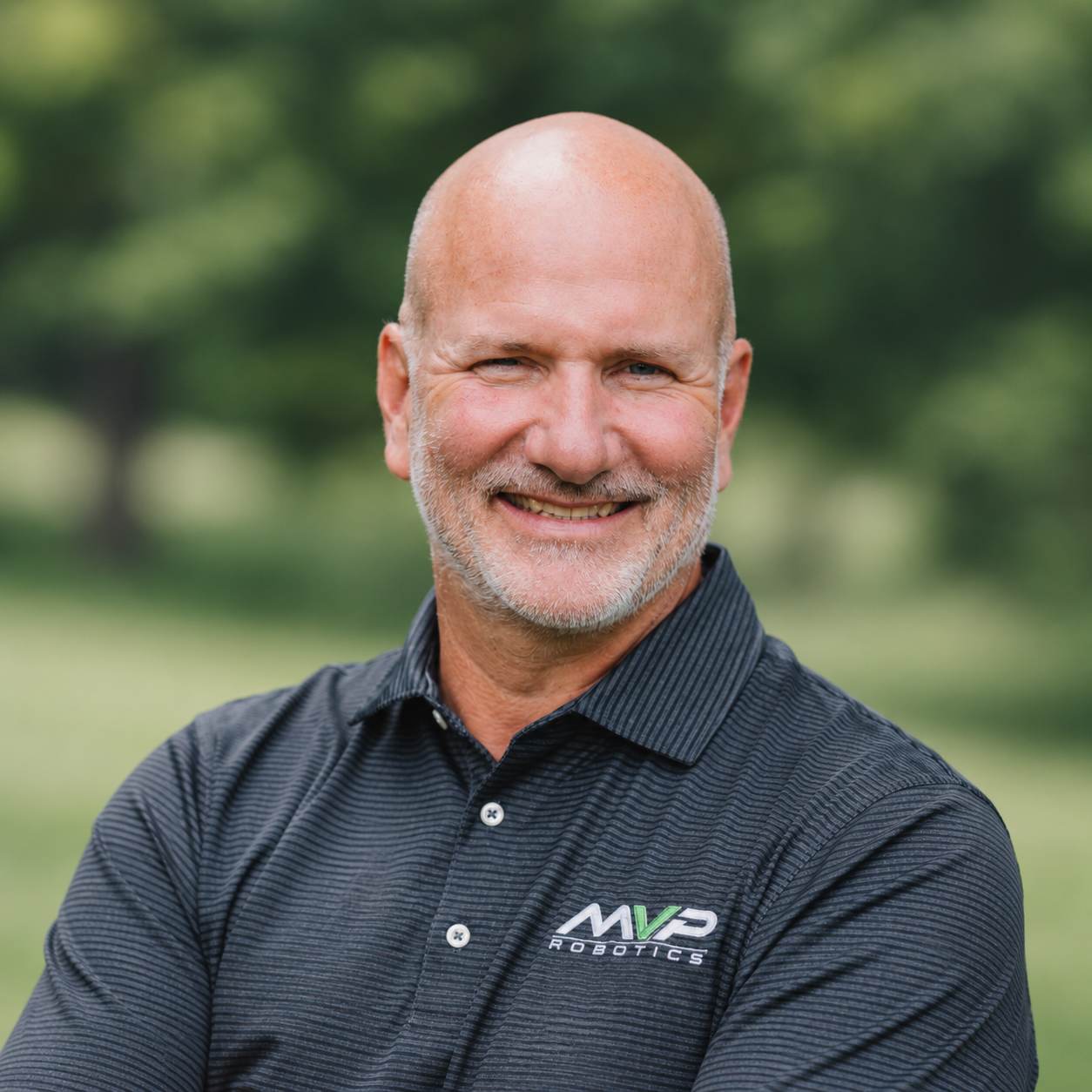Smiling bald man in a dark polo shirt outdoors with blurred green trees in the background