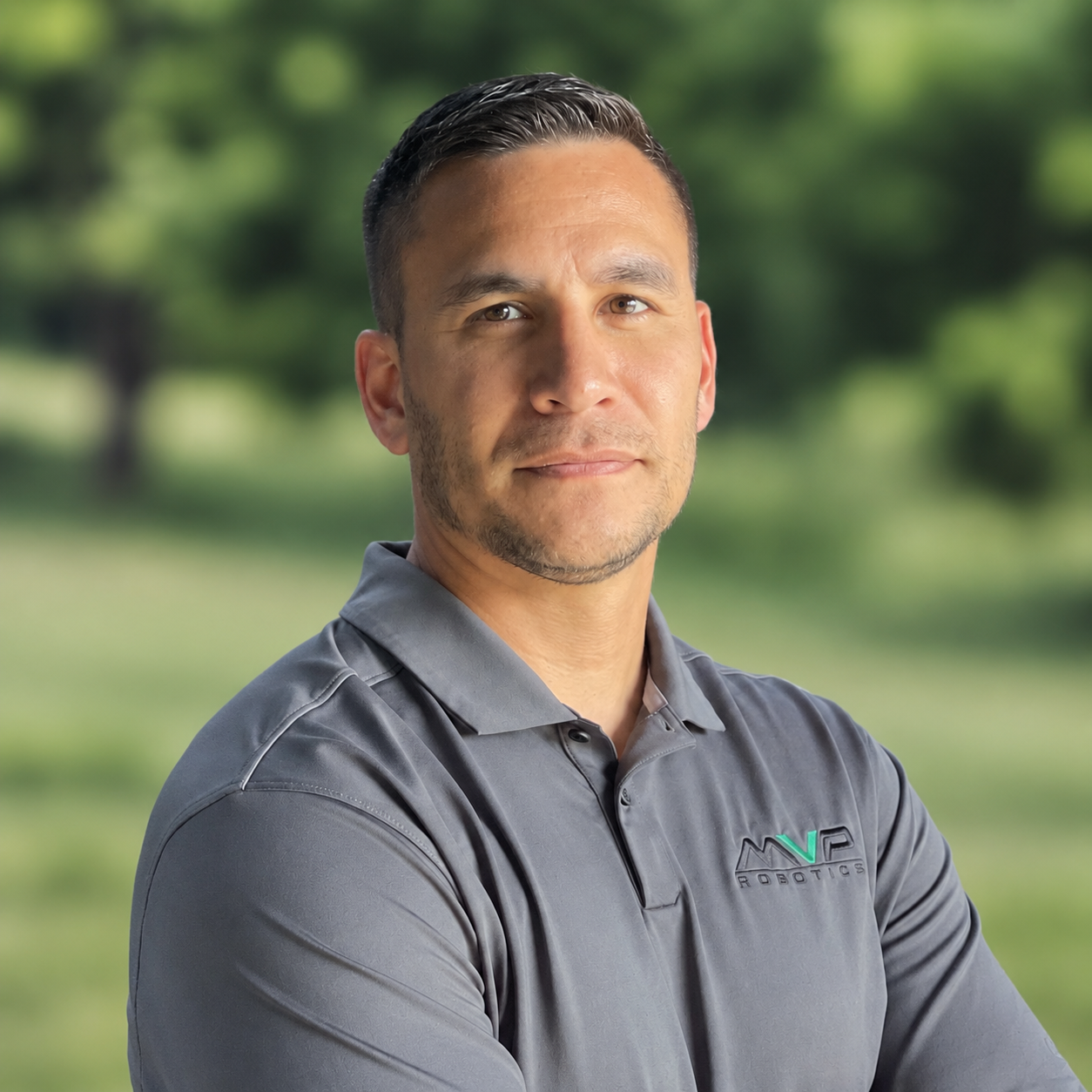Portrait of a man in a gray polo, standing outdoors with arms crossed and a blurred green background
