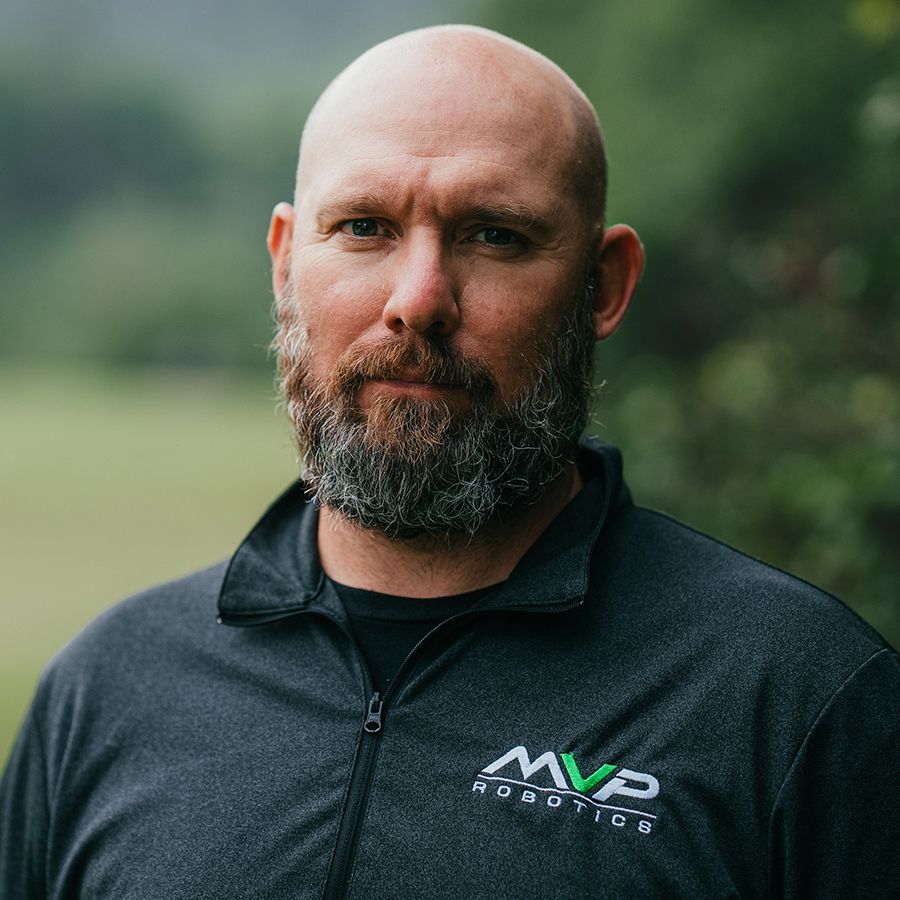 Close-up portrait of a bald man with a beard in a black zip-up jacket outdoors.