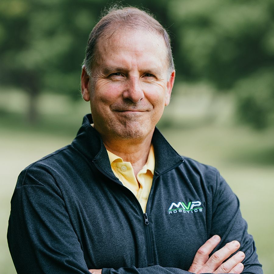 Smiling man in a dark jacket with arms crossed outdoors, wearing a yellow shirt