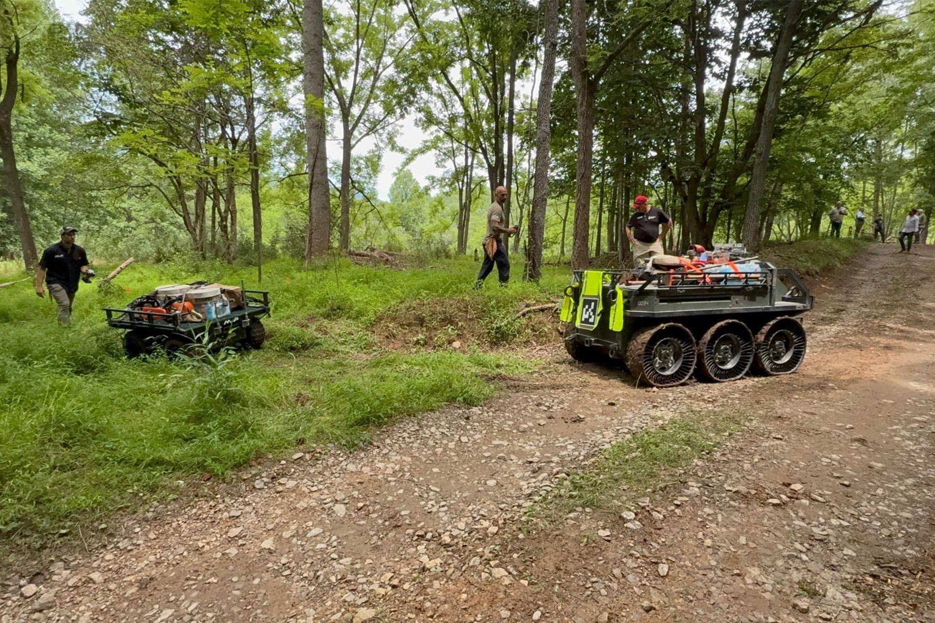 Small tracked vehicle and ATV on a dirt trail in a wooded park, with people nearby