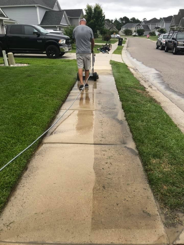 Person pushing a lawn mower on a wet suburban sidewalk beside green lawns and houses.