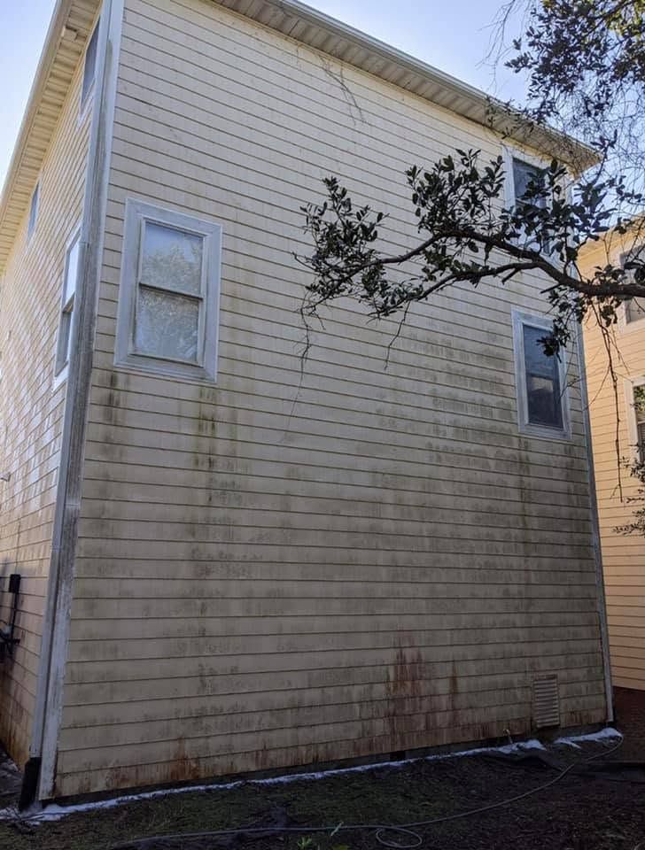 Side view of a beige house with two windows, weathered siding, and a tree branch overhead.